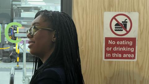 Kemi Badenoch stands by a door with a sign reading: "Not eating or drinking in this area"