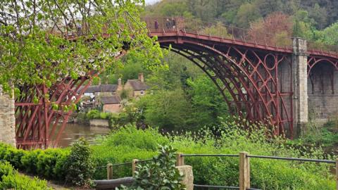 A rust coloured bridge over a river. People can be seen walking on the bridge. In the foreground trees, bushes and a path can be seen