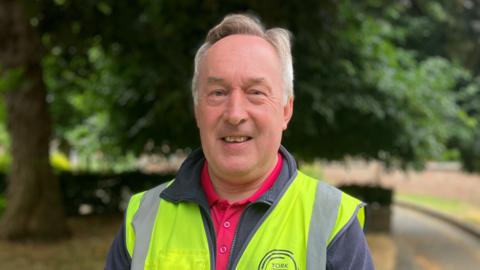 A man with short grey hair in a hi-vis vest smiles at the camera.