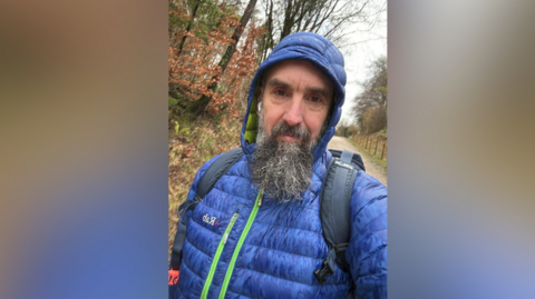 A bearded man in a blue puffa jacket with the hood up looks at the camera. He is wearing a backpack on a woodland path