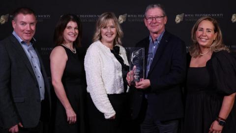 Five people are standing together smiling. Included is Environmental correspondent Louise Cullen, Newsline presenter Tara Mills, Damien Magee (TV news editor),holding an award and TV director Cara. 