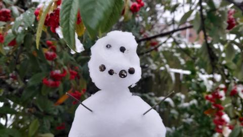 A small snowman has two stick arms and a smile made from whole peppercorns. In the background, there is a light dusting of snow on a bush.