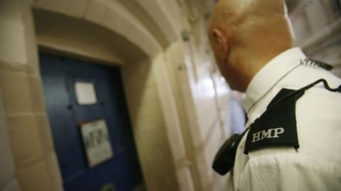 A prison guard in a white shirt with a walkie-talkie over his left shoulder stands in the foreground looking at a blue cell door 