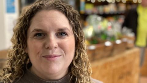 A woman with long, curly, light-brown hair stands in front of the counter at a community grocery shop.