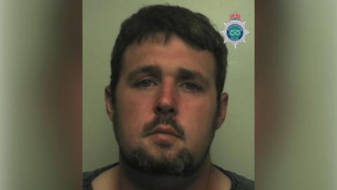 Robert Gillan, a man with dark brown hair and a beard, is standing against a light-coloured backdrop. There is a Staffordshire Police emblem in the top right hand corner of the image.