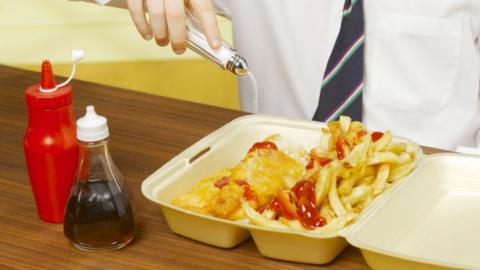 A boy wearing a white shirt and blue tie pours salt on a box of fish and chips, which is doused in ketchup. A bottle of ketchup and vinegar sits next to it on a table.