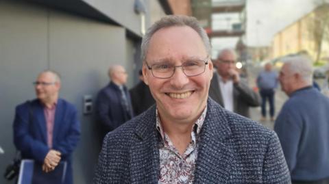 A man with glasses, a paisley shirt and a tweed coat.