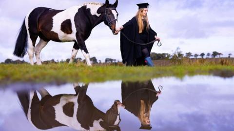 Woman in graduation gown leads a brown and white horse, both reflected in water