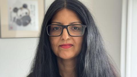 A head shot of a woman with long black hair, red lipstick and black square-rimmed glasses.