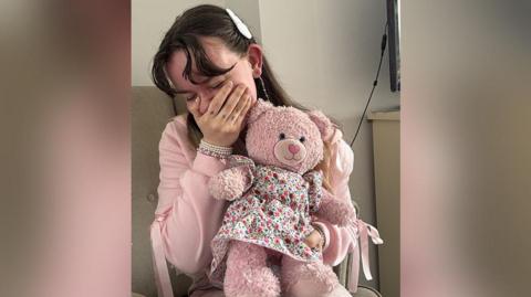 Lyric Holmes, with long brown hair wearing a pink top has her hand over her mouth and crying with happiness. She holding a pink teddy bear that is wearing a floral dress.