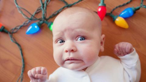 A generic image of a baby in a white onesie, lying in amongst a string of Christmas lights, with a comical worried expression on its face.