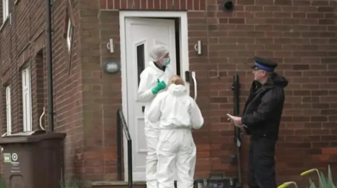 Policeman in black uniform and cap speaks to two forensic officers in white scrubs as they stand outside the entrance to a brick house.