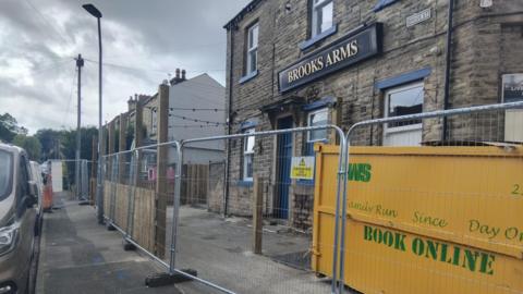 A pub behind a series of metal fences with a yellow skip to the right