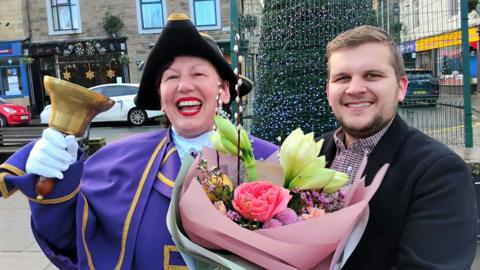 A smiling woman in a traditional town crier costume including a black tricorne hat and purple robe, holding a large golden bell. She is being presented with a large bouquet of flowers by a man in a black jacket and chequered shirt. 