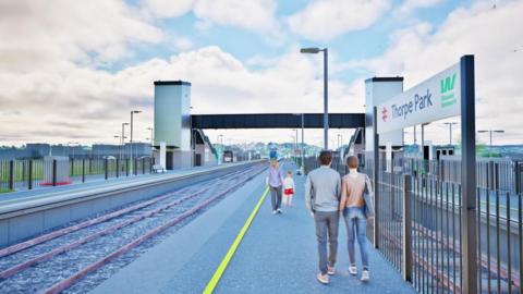 Computer-generated image of the proposed Thorpe Park Rail Station showing two railway platforms, a footbridge with lift towers, railway tracks, safety fencing, platform lighting and people walking along the platform beside a station sign.