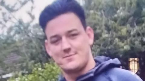 Macaulay Branighan stands in front of some trees and a slight smile on his face. He is clean shaven with thick black hair swept back. The image is quite blurry. 