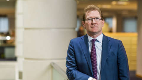 Dr Andrew McLaughlin wearing a blue suit jacket and a red and white spotted tie. He is stood to the right leant against a railing. 