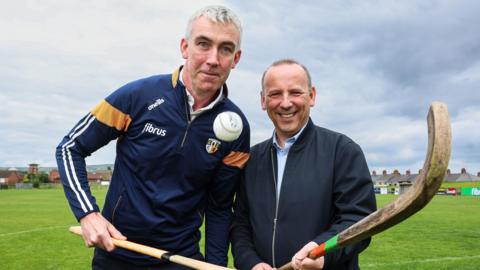 Antrim GAA chairperson Seamus McMullan wears a blue sports jacket and Rev David Moore a navy zip-up jacket. The pair are holding a wooden shinty stick and hurling stick on a green pitch. 