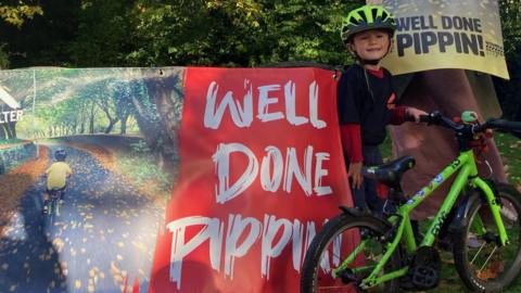 Pippin wearing a helmet, standing with his bike in front of banners that read "well done Pippin"