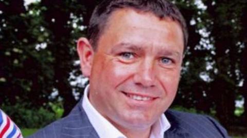 Jesse Richards pictured in a navy blue pinstripe jacket and white shirt. He had short brown hair and was clean shaven.