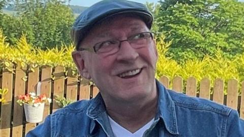 A man wearing a cap, a denim shirt and rectangular glasses sitting in a garden - he is smiling and looking off to the right of the camera