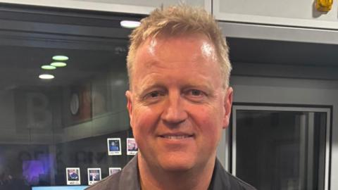 A photograph of a man wearing a black shirt looking directly into the camera. There is a glass window and door behind him.