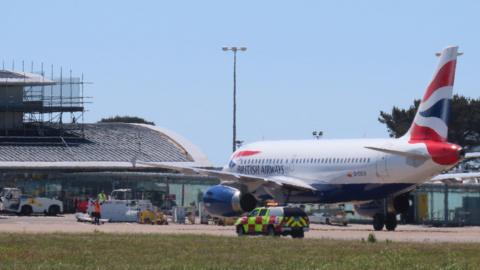 A view of a white red and blue British Airways aircraft in front of the airp[ort terminal building in Guernsey.