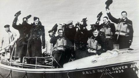 A black and white photo of the RNLI crews from WW2. They are in a lifeboat raising their hats in the air to wave. 