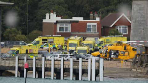 Ambulances, emergency response vehicles and an air ambulance helicopter are gathered near the seafront during an emergency operation.