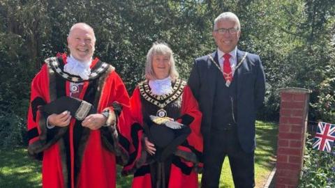 Two men and a woman are standing in a garden. The man and woman on the left are wearing ceremonial robes and  the man on the right is wearing a suit and ceremonial chain.