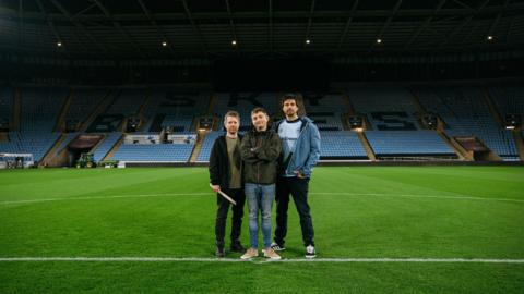 Three men standing in the middle of a football pitch. The back of the stadium can be seen with blue seats lined across the photo. 