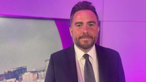 Portrait of a person wearing a dark business suit, white shirt and patterned tie, standing indoors in front of a purple-lit backdrop. A cityscape image is partially visible on the left, while the background features modern wall panels illuminated with pink and purple lighting. The person is centred in the frame and facing the camera.
