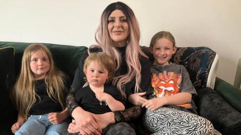 A woman is sitting on a sofa with a small boy on her lap and two young girls sitting either side of her. 