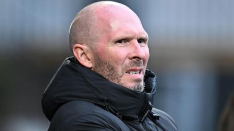 Michael Appleton is wearing his black puffer coat watching on from the sidelines during a match with a worried look on his face