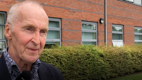 Kevin Townsend wearing a blur floral shirt and dark jacket standing in front of a hedge, in the background there is the red brick school building.