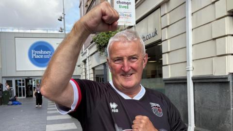 A man in his 60s with short grey hair fist pumps the air in celebration. He is wearing a black and white Grimsby Town shirt. 