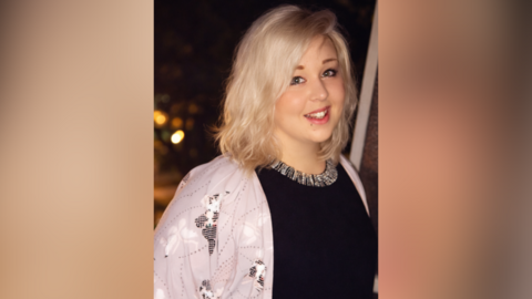 A woman is wearing a pink jacket and a black top, with a necklace. She has blonde hair and is smiling at the camera.