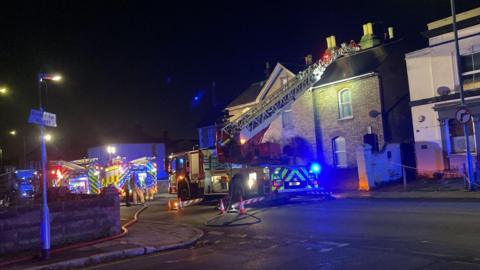 Three fire engines outside a row of terraced houses on a main road at night. One of the engines has an aerial ladder which has been extended up to the roof of the houses