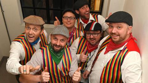 Six members of the Lancashire Hotpots stand on a staircase in rainbow waistcoats and flat caps. They are grinning and have their thumbs up.