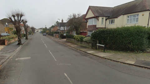 A residential tree-lined street with houses on either side and a car is parked on the right-hand side. 