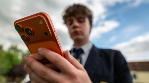 A teenage boy wearing a navy school uniform blazer, tie and white shirt holds an orange smartphone out in front of him.