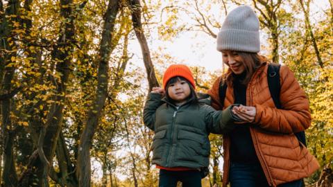 A woman and her child walk through a forest, the leaves are orange and autumnal.