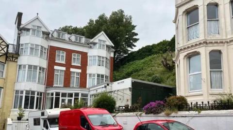 A four-storey, red-brick building with large bay windows on either side, flanked by a modern, single-storey building which juts out on the right - a steep green hill rises behind it