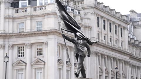 A grey statue of a man wearing a suit, marching forward, carrying a flag that covers his face