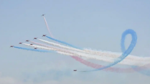 Nine aeroplanes in a line formation with two twirling in the sky with white blue and red smoke coming out behind them.