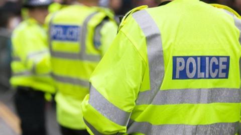 Three police officers standing outside, all with their backs to the camera. They are all wearing fluorescent yellow jackets with silver stripes across the backs and sleeves. On the back of the jackets there is a blue rectangle with the word, POLICE written inside.