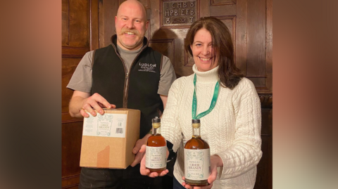 A man and a woman stand next to each other in a room with wooden panelled walls. The man is bald with a blond moustache and is holding a wooden box. The woman has long brown hair and she is holding two bottles of brown liquid. 