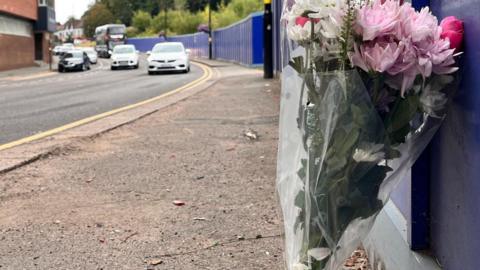 A blunch of pink and white flowers lean against a dark blue hoarding. The hoarding is on one side of busy one-way road.