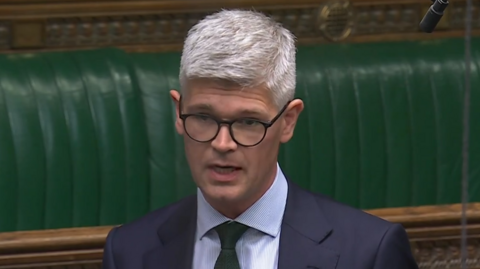 A man with short white hair speaking in the House of Commons. He has black glasses, a green tie and a navy suit on. He is standing in front of green leather benches.
