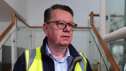 A dark haired bespectacled man in a high vis jack and open necked shirt